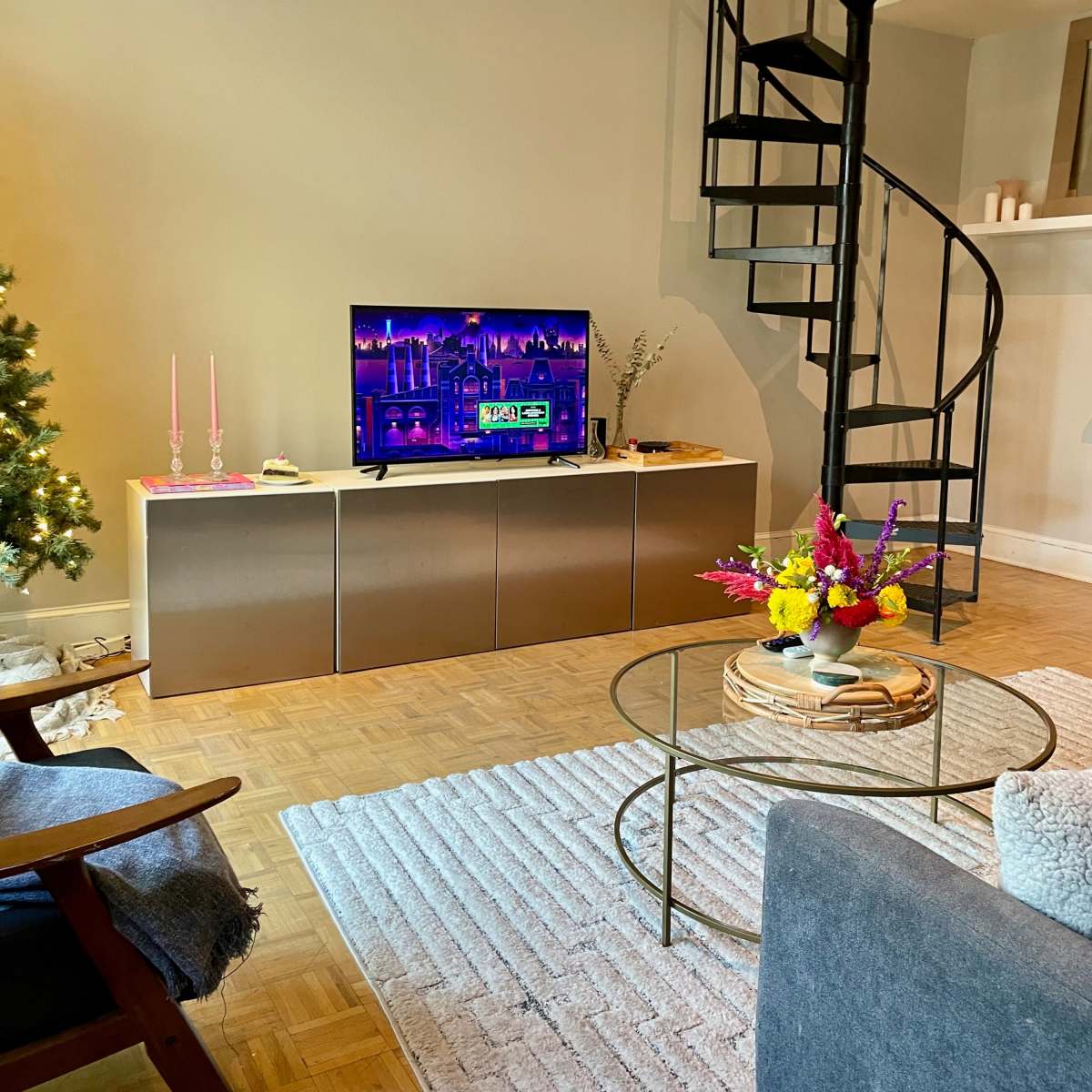 A cozy living room with a television on a cabinet, a spiral staircase in the background, a small coffee table with flowers, and a decorative rug on the wooden floor.