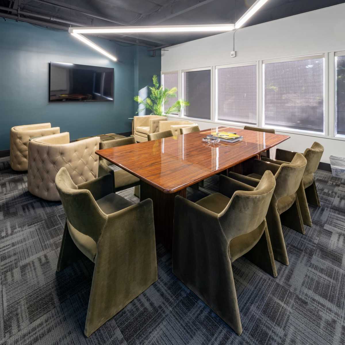 A modern conference room features a large wooden table surrounded by upholstered chairs, with a TV on the wall and large windows allowing natural light.