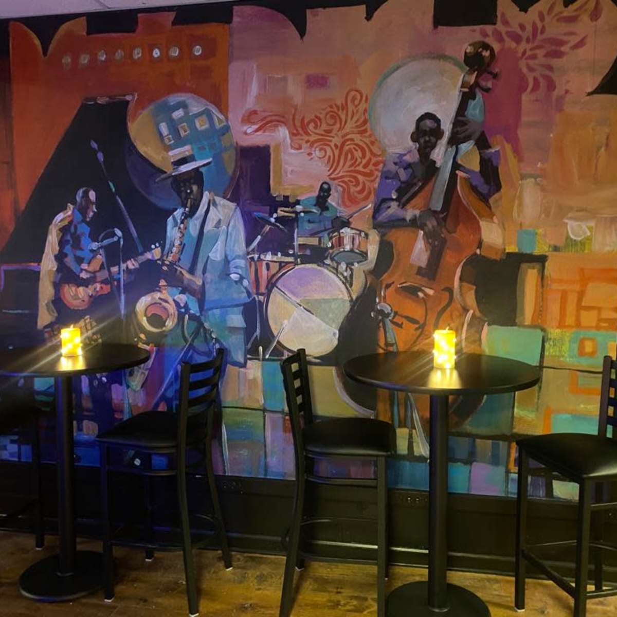 A vibrant mural of a jazz band on a wall behind empty black tables and chairs in a dimly lit venue.
