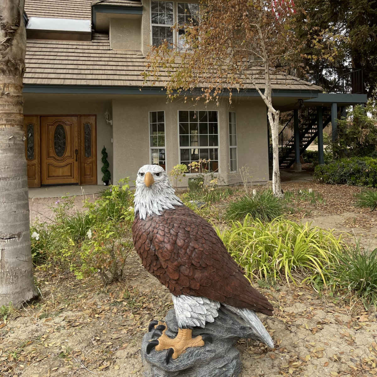 A large eagle statue stands in front of a two-story house with a staircase and an American flag hanging from the porch.