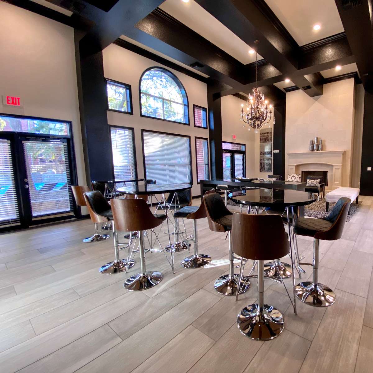 A modern lounge area featuring round tables with high-backed chairs, a large chandelier, and a fireplace under a high ceiling with exposed beams.