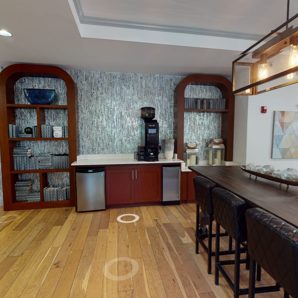 A modern kitchen area with wooden flooring, featuring a coffee machine on a countertop, shelves filled with towels, and a long dining table with black chairs.