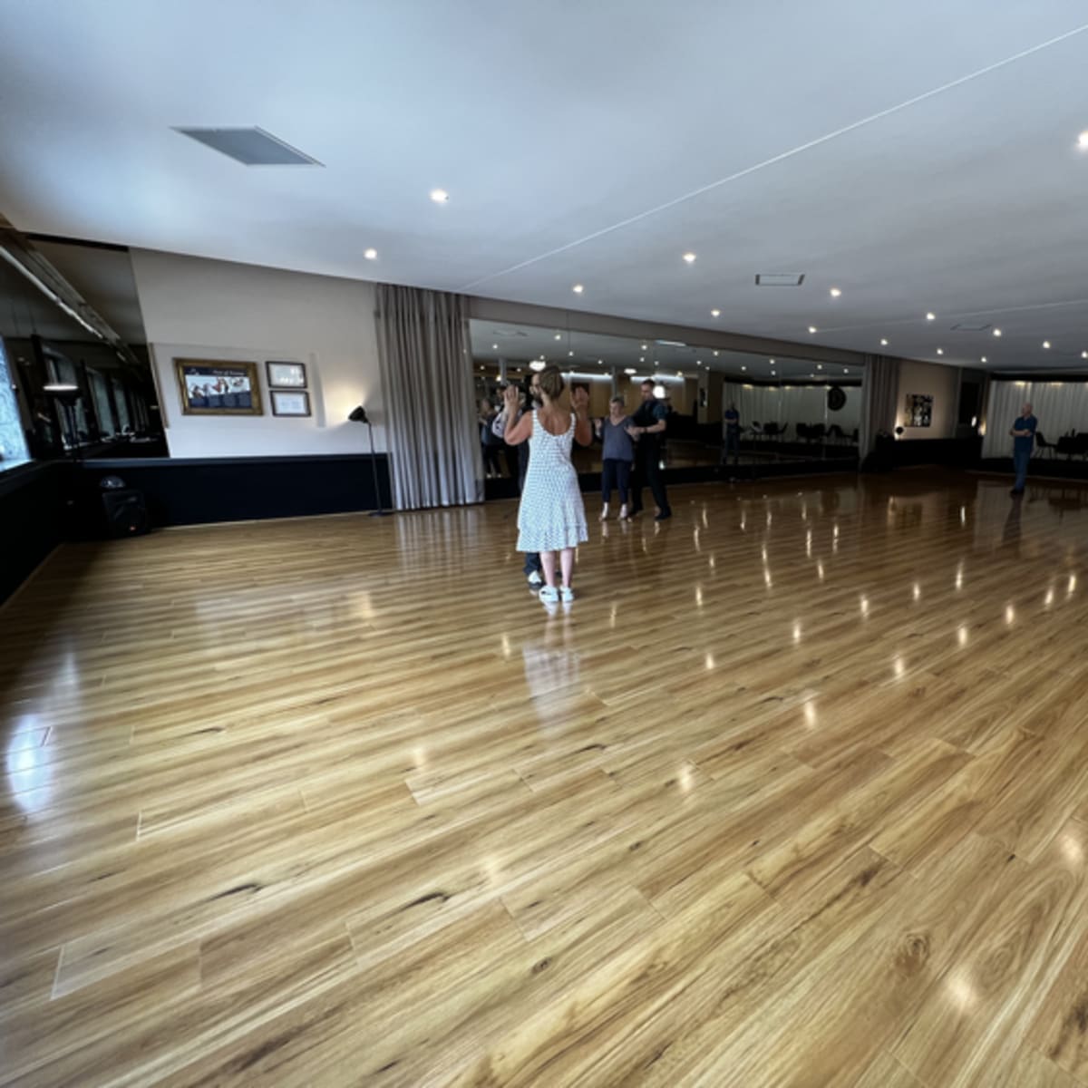 A spacious dance studio with polished wooden floors, mirrors on one wall, and attendees practicing in pairs.