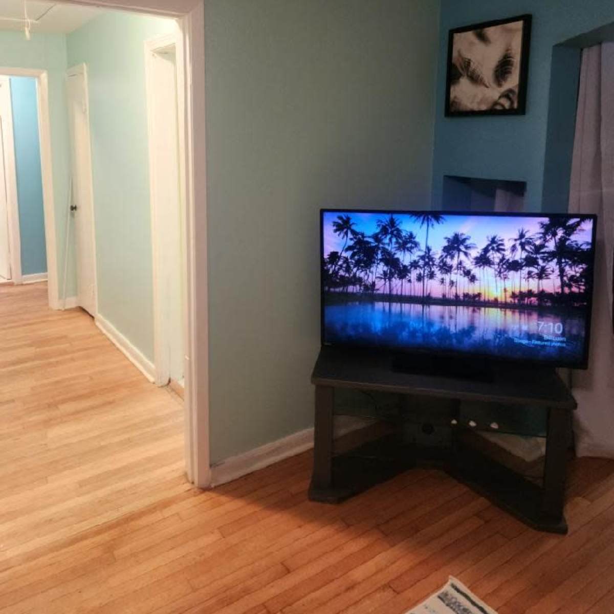 A living room with a television displaying a tropical sunset scene and a view of a hallway with a light blue wall and wooden flooring.