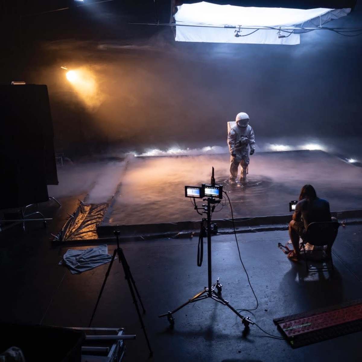 A person dressed in a spacesuit stands on a fog-covered floor while a photographer sits on a chair, capturing the scene.