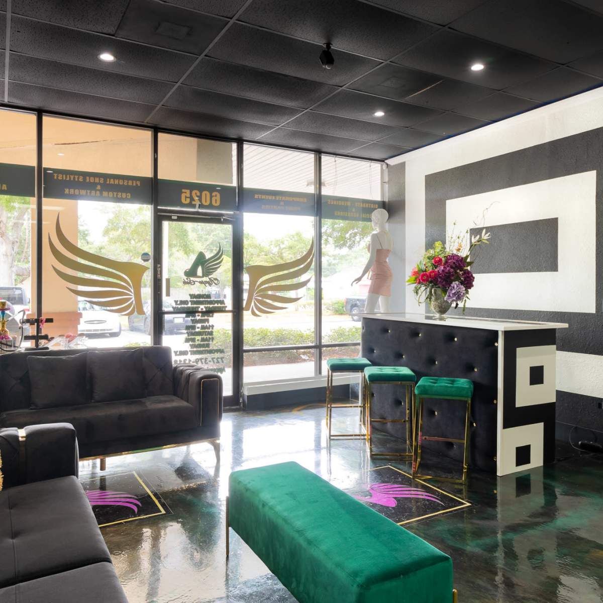 The interior of a modern salon features black sofas, a green bench, a stylish reception area with high stools, and large windows showcasing outdoor views.