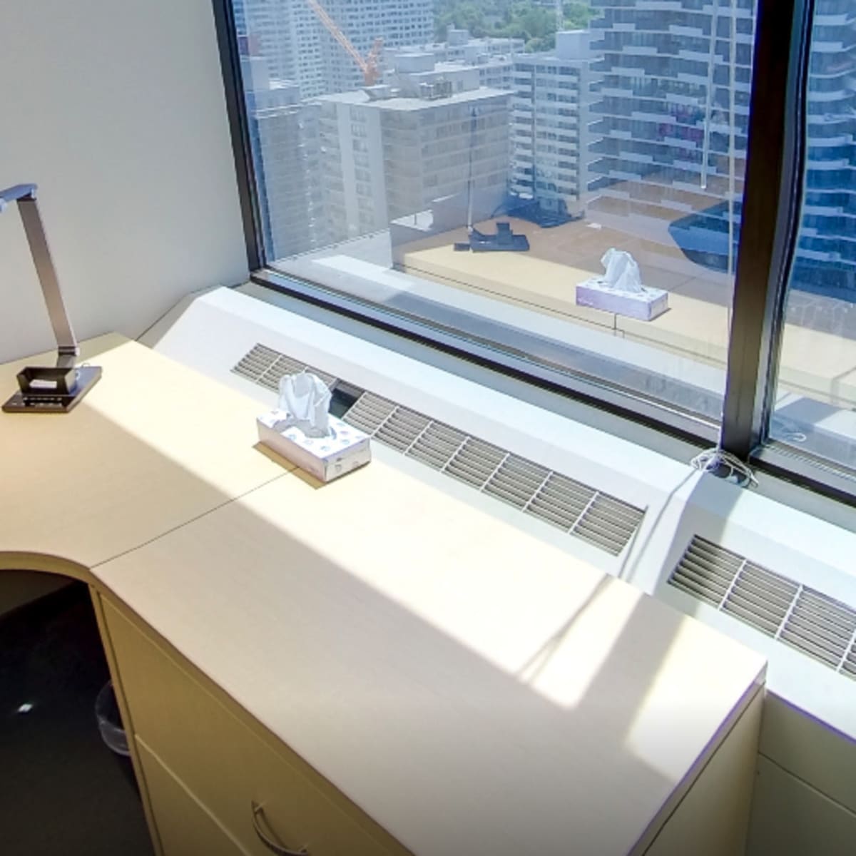 An office space with a desk, a telephone, and a lamp, along with a tissue box on the windowsill, overlooking a cityscape.
