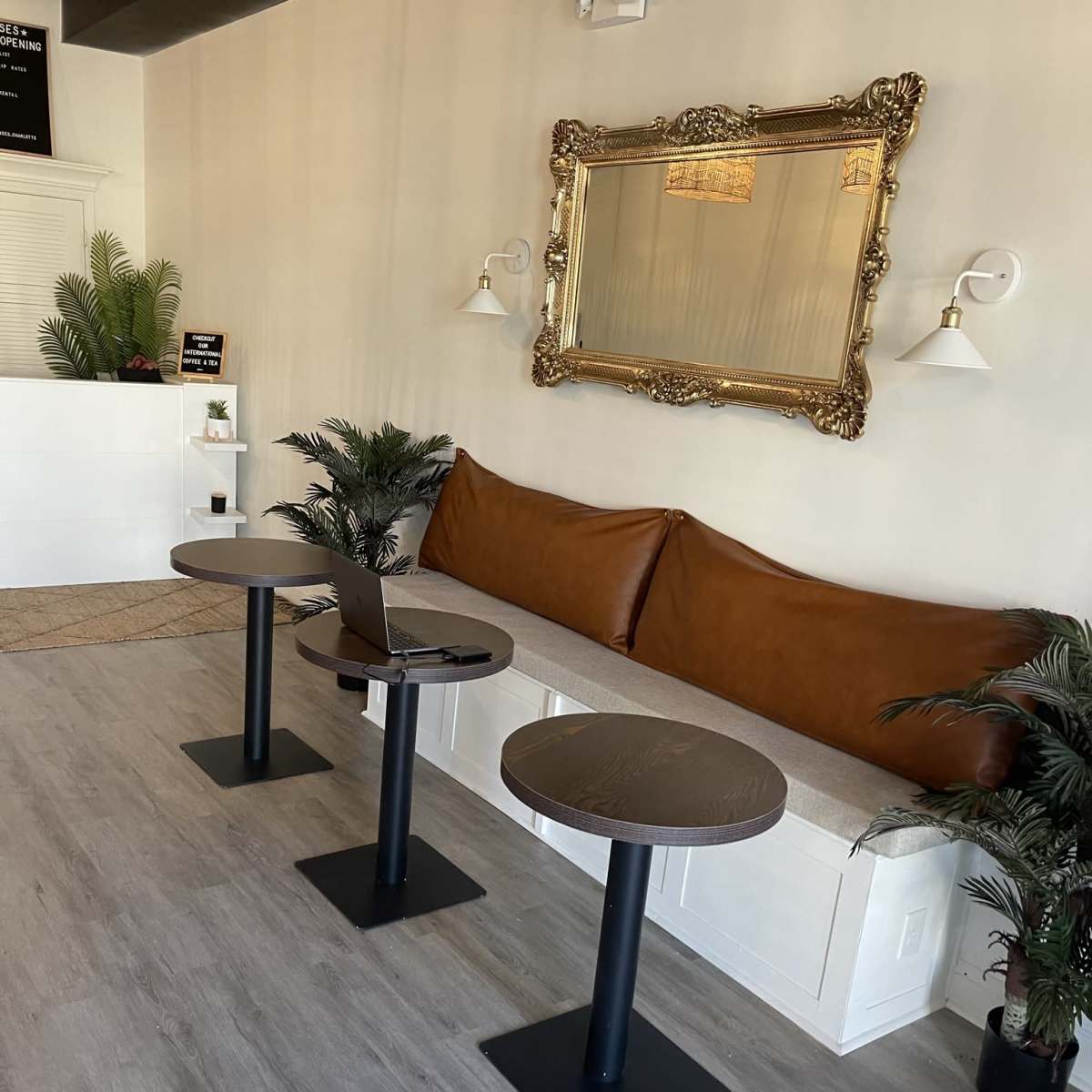 A modern interior space with three black tables, a brown leather bench, a large ornate mirror, and potted plants along the walls.