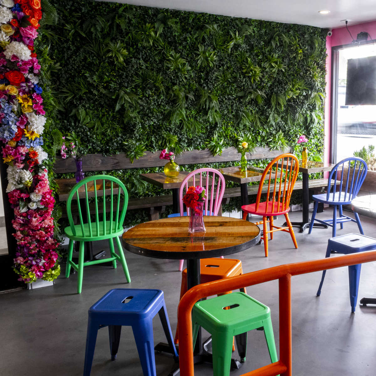 A vibrant café interior with colorful chairs and tables arranged around a lush, green wall adorned with flowers.