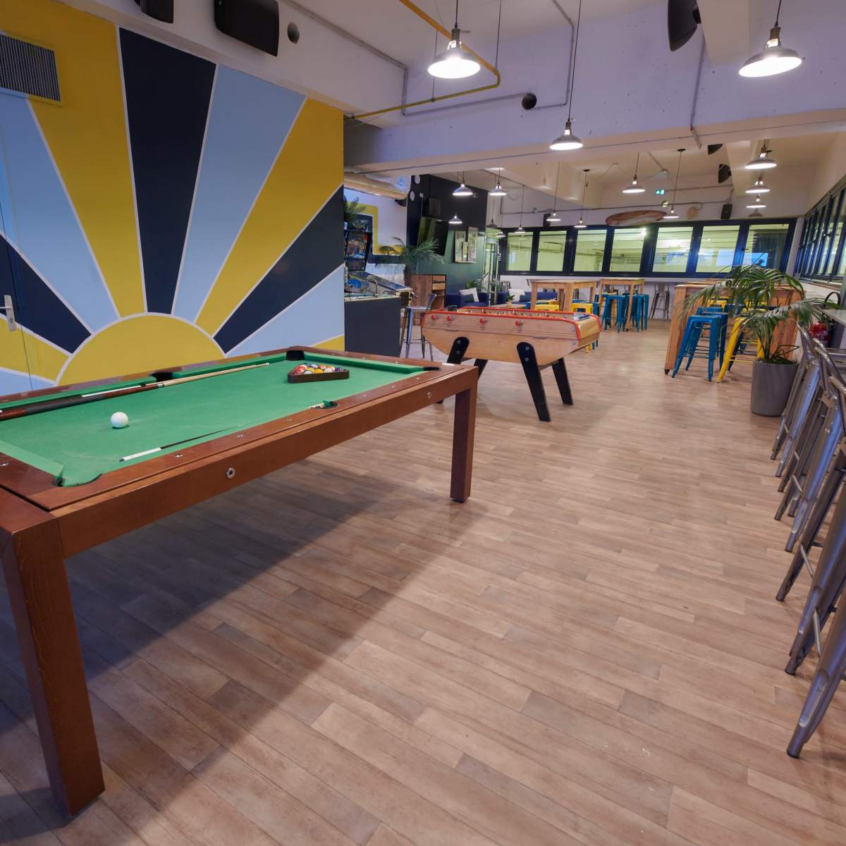 A recreational room with a green billiard table, colorful wall art, and seating arrangements, alongside foosball and dining tables.