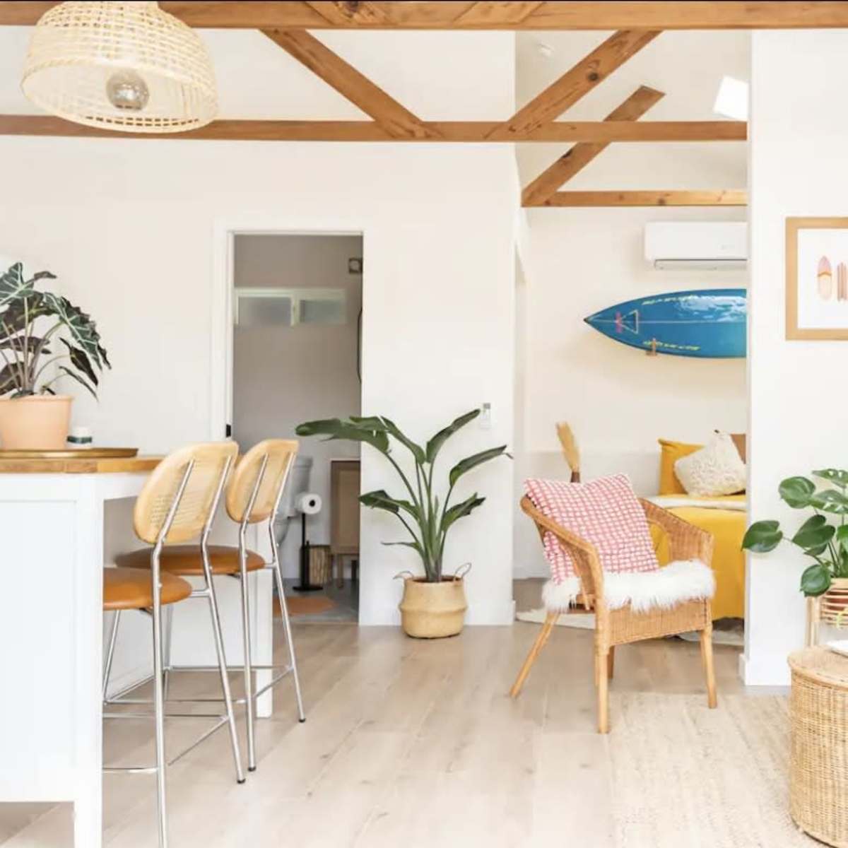 A bright, open living space with wooden beams, a kitchen island, a cozy seating area, and a surfboard on the wall.