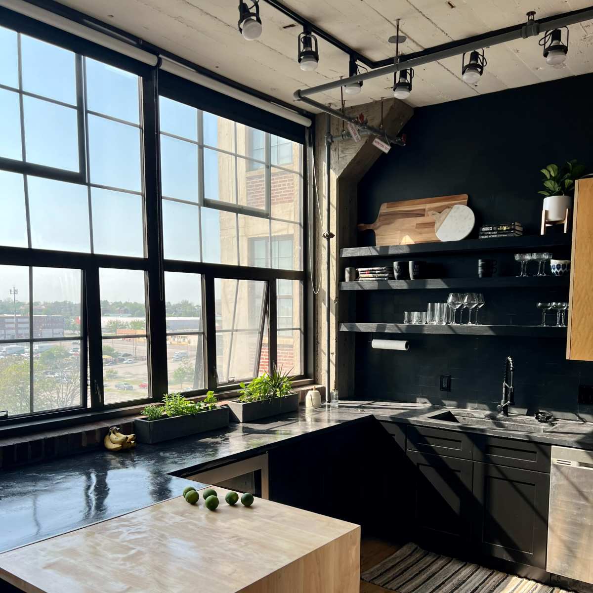 A modern kitchen featuring a large window, dark cabinetry, a minimalist countertop, and a wooden island with three limes placed on it.