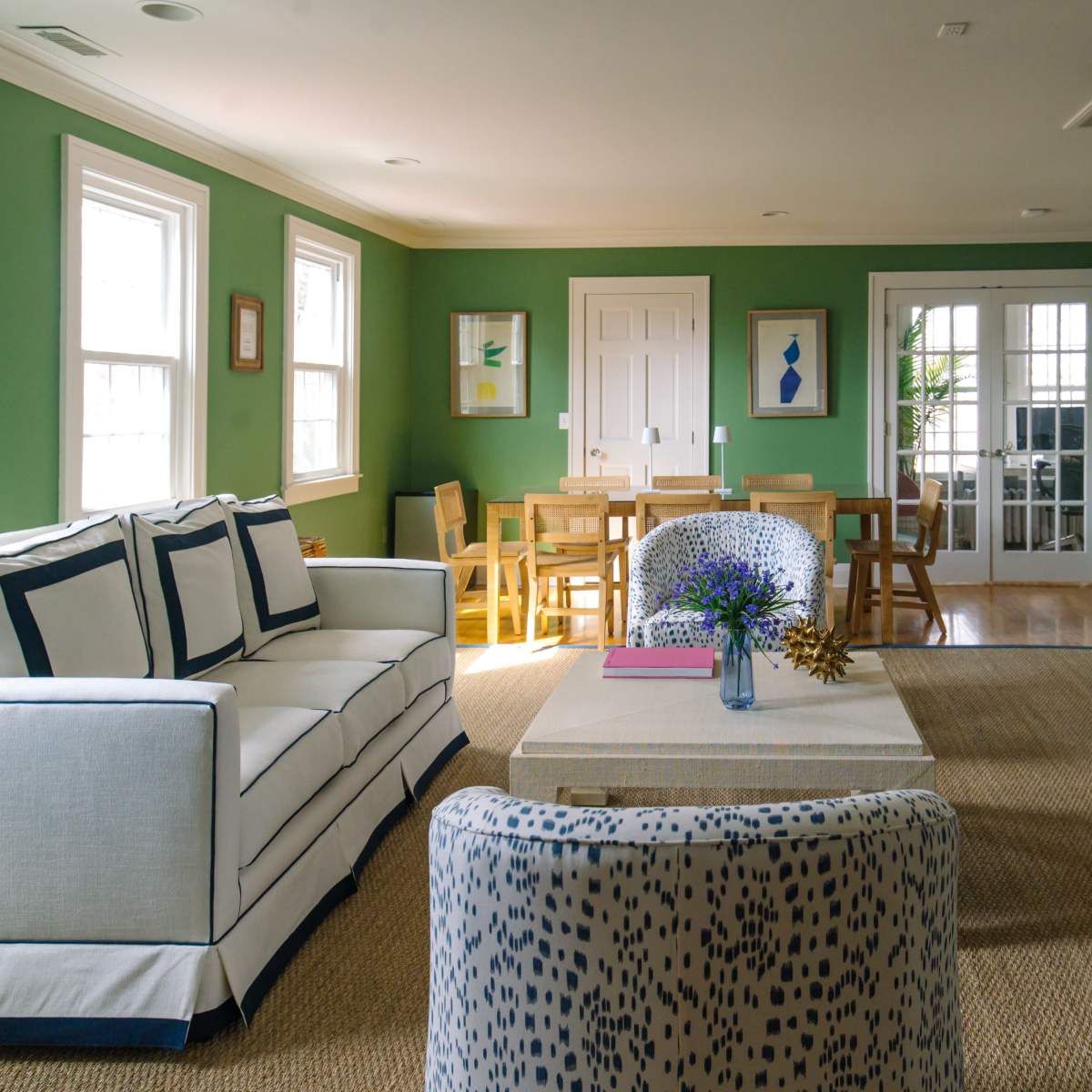 The room features green walls, a white sofa with navy trim, wooden dining chairs, and a glass door leading to an outdoor space.