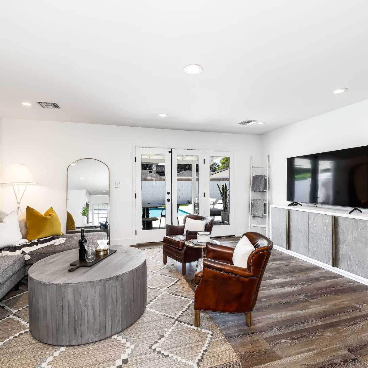 The living room features a gray sectional sofa, a circular coffee table, and a large television, with a view of a pool visible through glass doors.