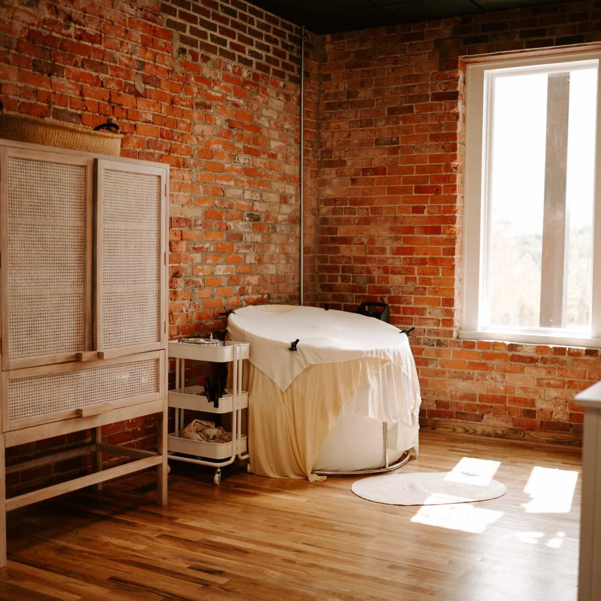 A room with exposed brick walls, a wooden floor, a large round massage table covered with a white sheet, and a window allowing natural light to enter.