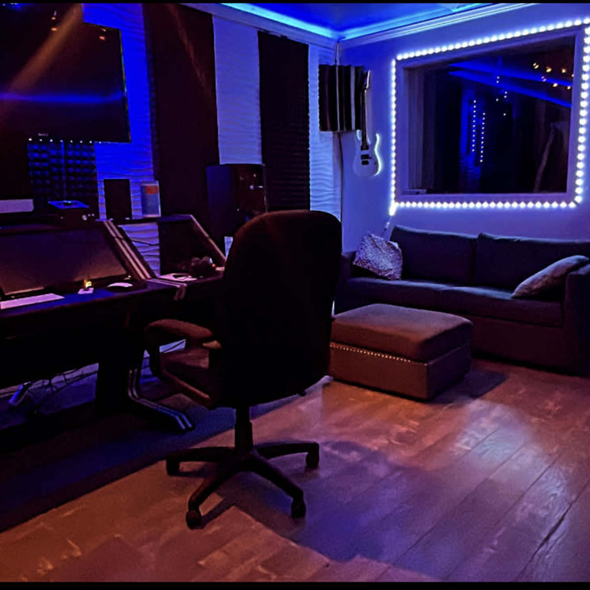 A music studio featuring a desk with computer equipment, a black swivel chair, a couch, and illuminated wall decorations.