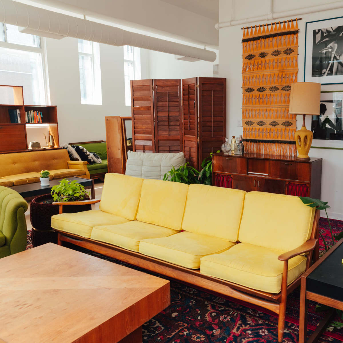 A brightly lit living room featuring a yellow couch, a wooden coffee table, and various decorative elements like plants and artwork.