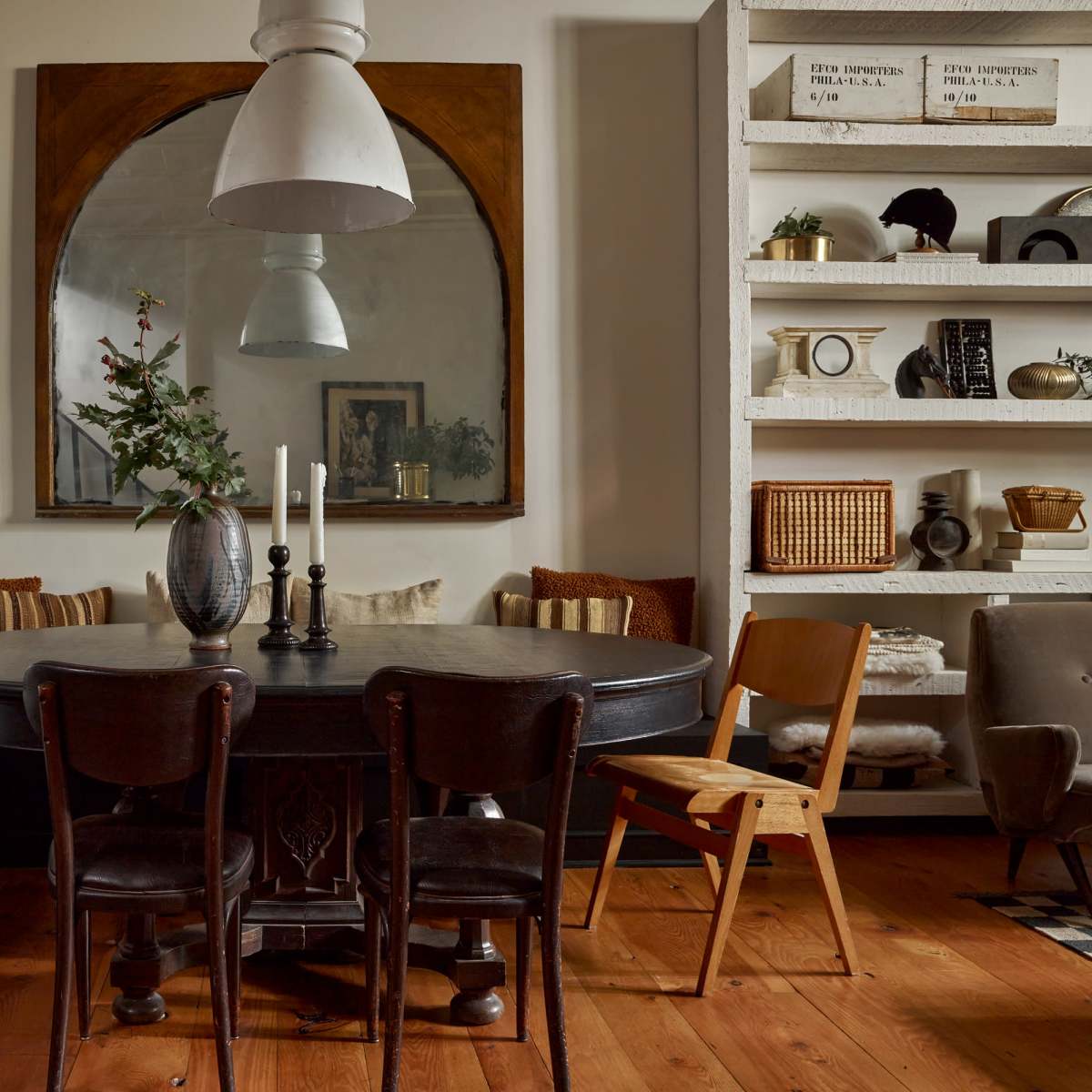 A circular black dining table surrounded by chairs is set in a cozy room featuring a large mirror and shelves filled with decorative items and plants.