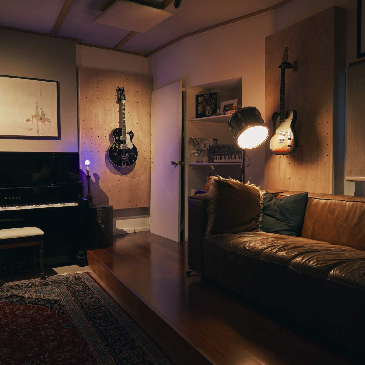 A music studio with a black piano, two guitars mounted on the wall, a brown leather couch, and a floor lamp illuminating the space.