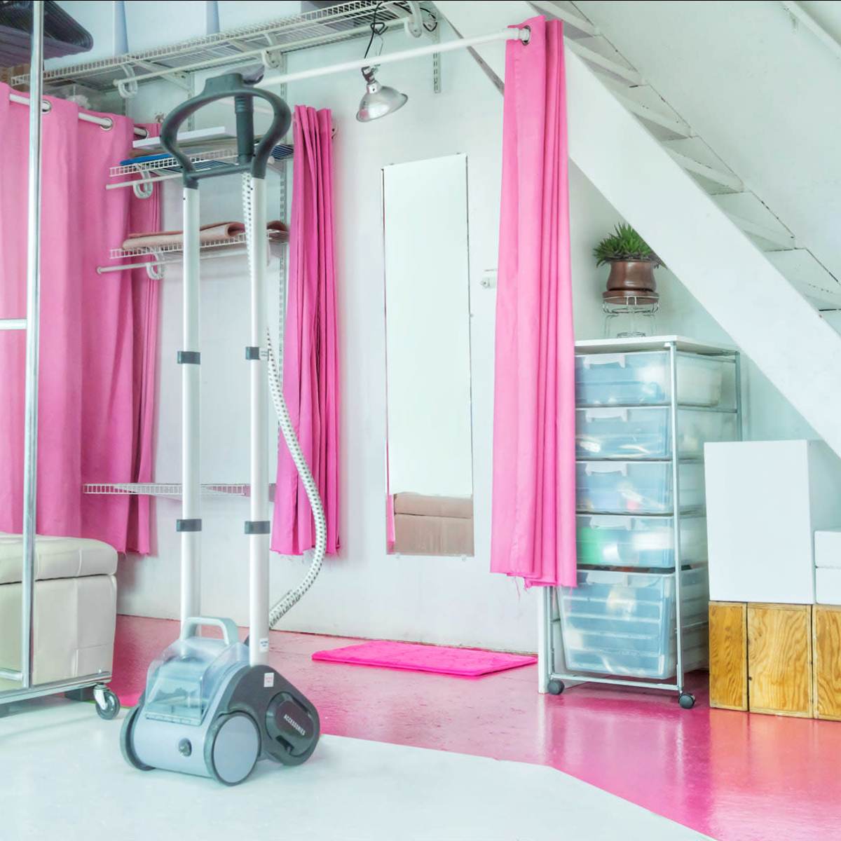 A bright, modern space featuring a white couch, pink curtains, a steamer, and storage units near a staircase.