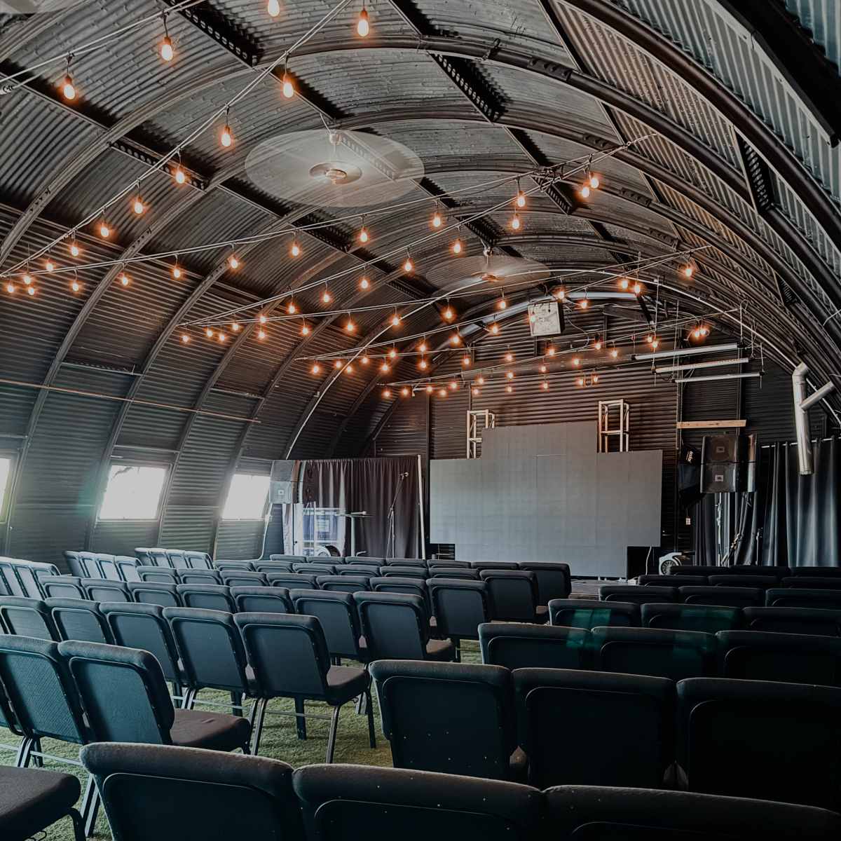 An indoor venue with rows of black chairs facing a stage, accented by string lights along the curved metal ceiling.