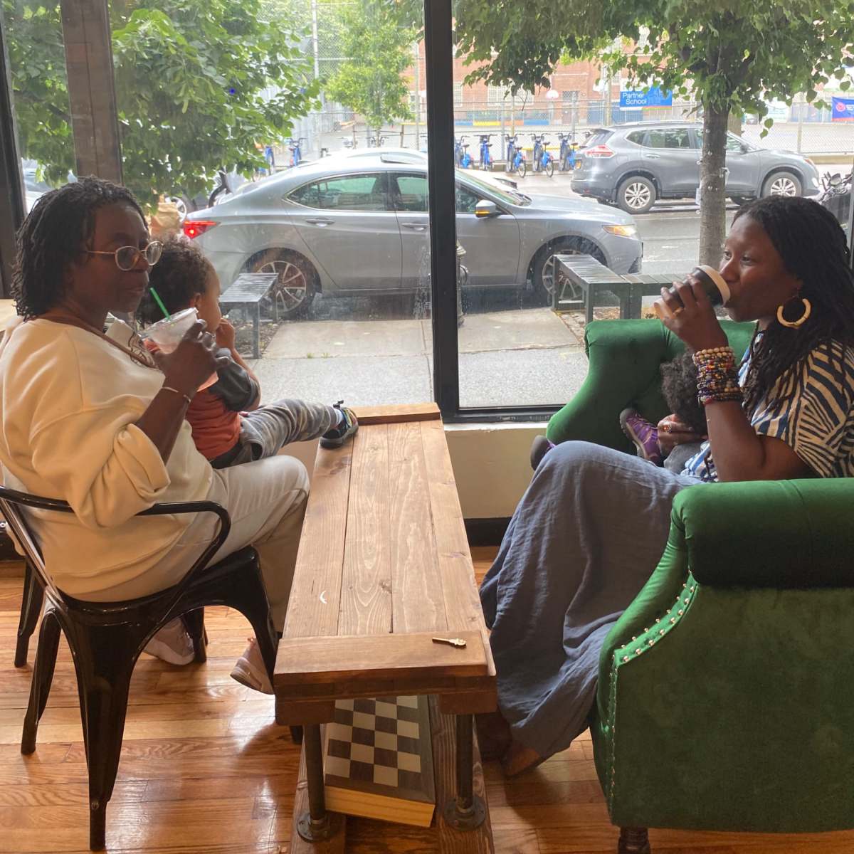 Two women sit at a table inside a café, one holding a child, while the other drinks from a cup, with cars and trees visible through the window behind them.