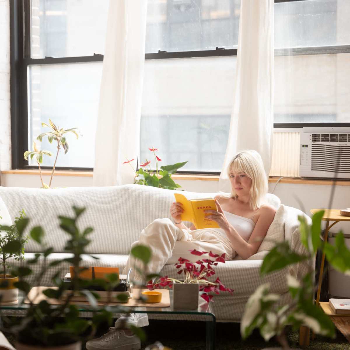 A woman sits on a white couch reading a book in a sunlit room filled with plants.
