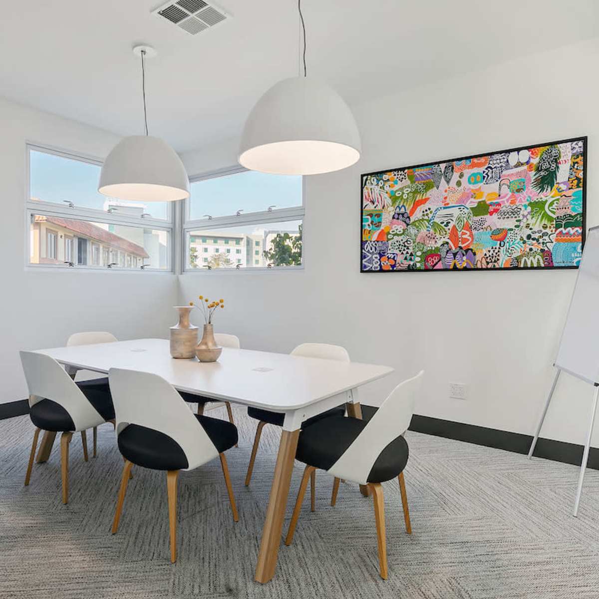 A modern conference room features a white table surrounded by six chairs, with large pendant lights, a colorful artwork on the wall, and a plant in the corner.