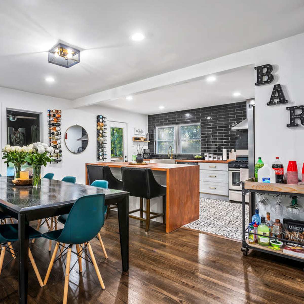 A modern kitchen and dining area featuring a wooden table with blue chairs, a central island with bar stools, and a bar cart along the wall.