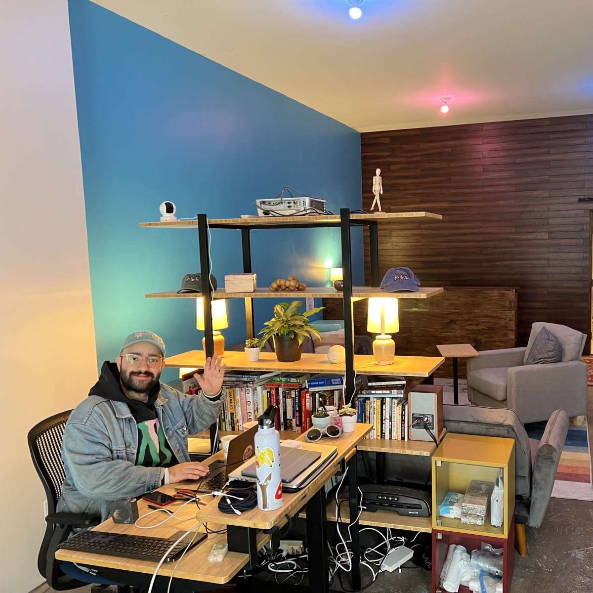 A man sits at a desk with a laptop in a modern office space, featuring a bookshelf and a seating area with a rug.