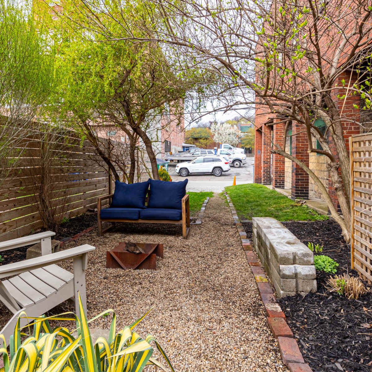 A gravel pathway leads to a seating area with a wooden couch and a fire pit, framed by low shrubbery and wooden fences.