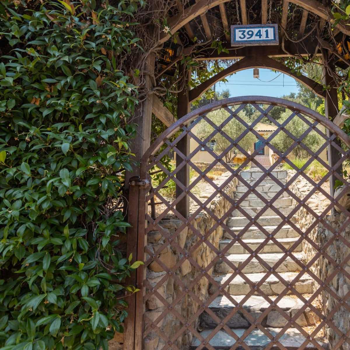 A wrought iron gate with a diamond pattern leads into a stone pathway bordered by greenery, with a house visible in the background.