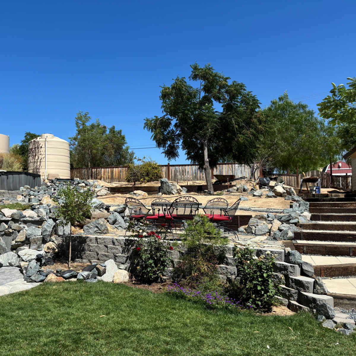 A landscape with a stone pathway leading to a rocky area, surrounded by greenery and a few trees, with an umbrella-covered seating area in the foreground.