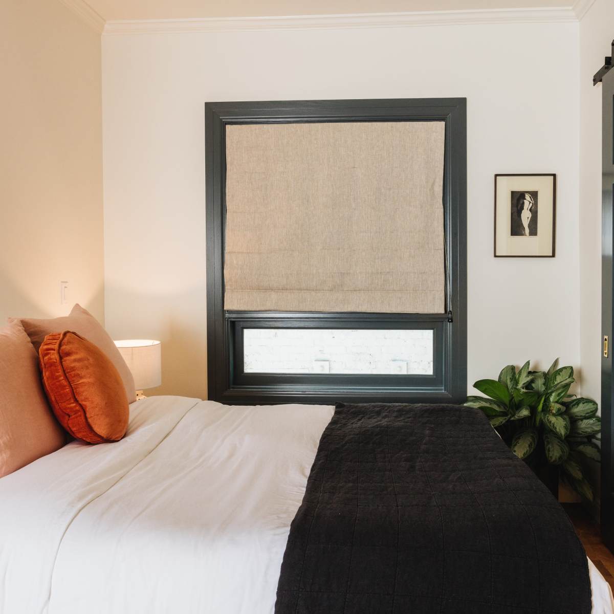 A neatly arranged bedroom features a bed with white and black bedding, two orange cushions, a framed picture on the wall, a potted plant, and a window covered with a beige shade.