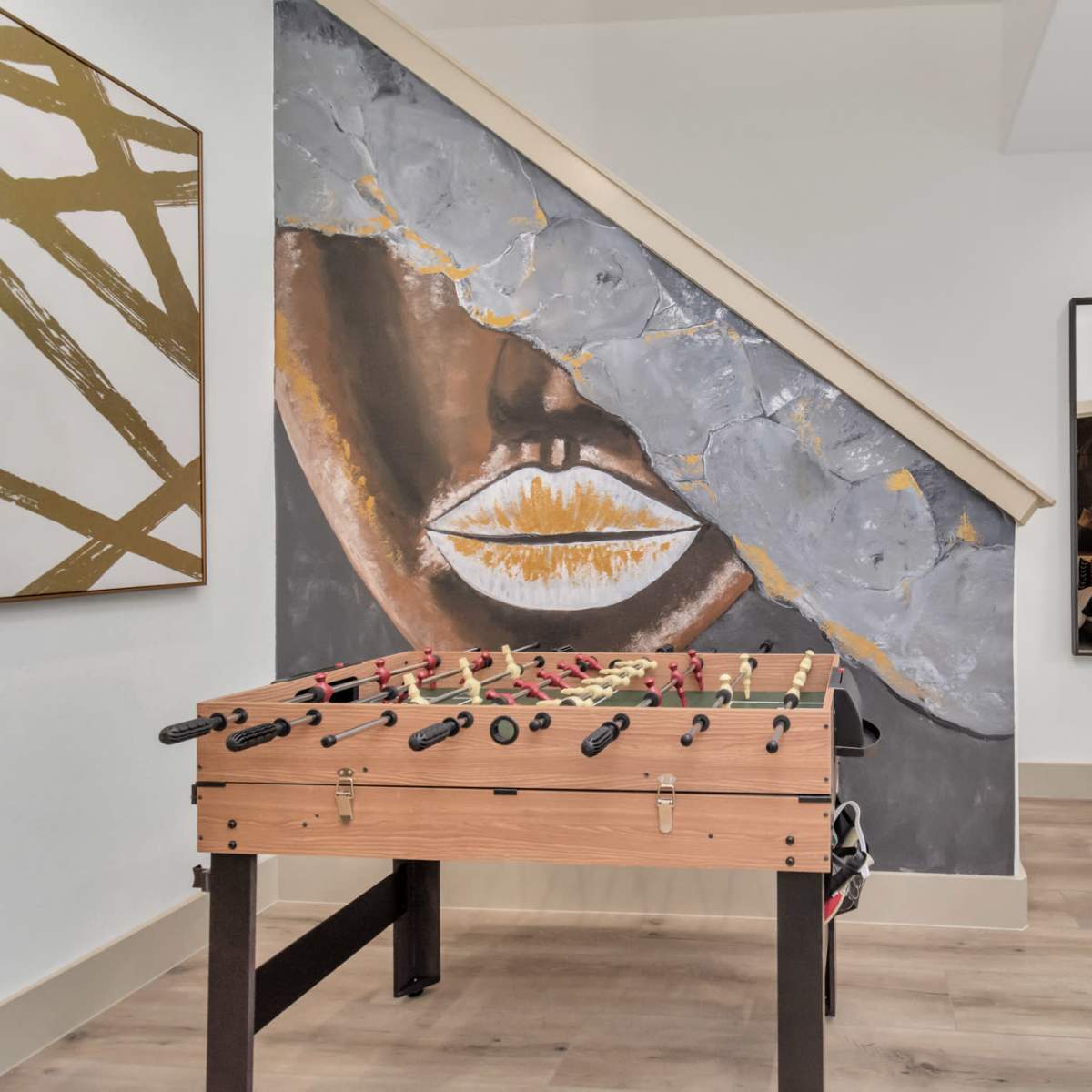 A foosball table sits near a staircase, accompanied by a large wall mural and a mirror in a well-lit room.