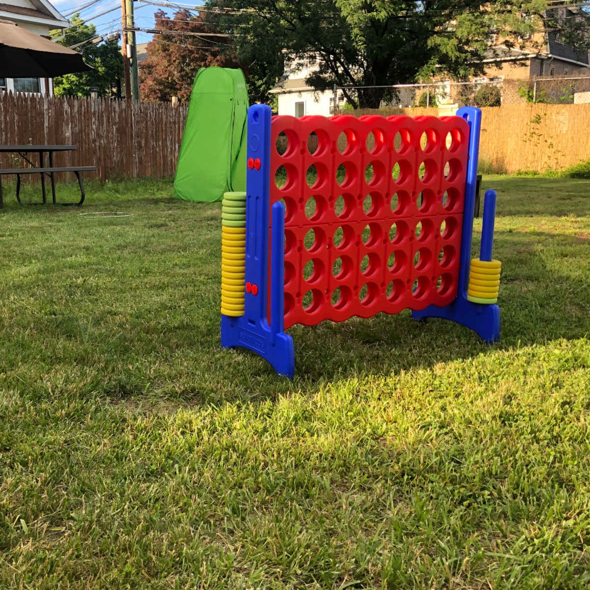 A large outdoor game of connect four is set up on a grassy lawn, with a picnic table and a green camping tent in the background.