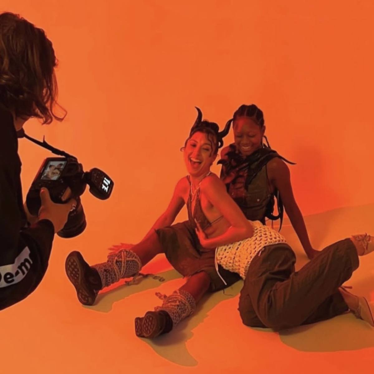 Two models pose playfully on a bright orange background while a photographer captures the moment.