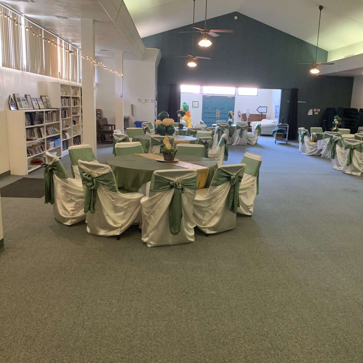 The room features multiple tables set with green tablecloths and white chair covers, with a library shelf visible in the background.