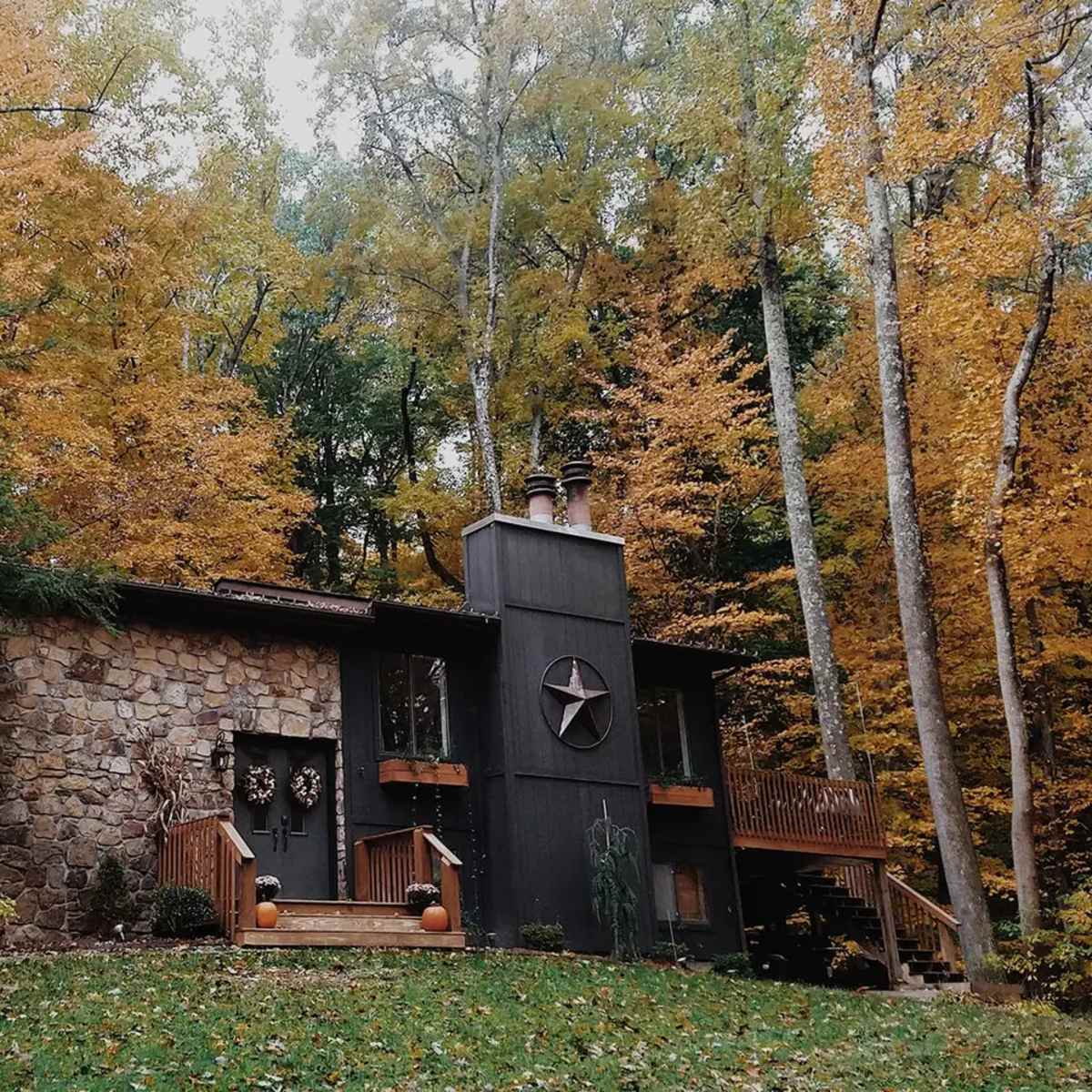 A stone and wooden cabin is surrounded by trees with autumn foliage, featuring a porch and a star decoration on the front.