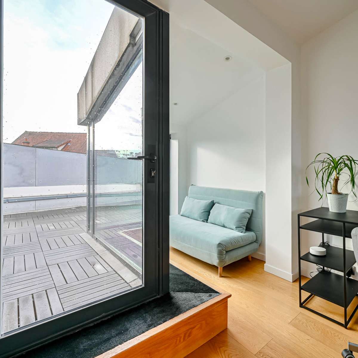 A small, modern room features a sliding glass door opening onto a terrace, a light blue sofa, and a minimalist black desk with a plant.