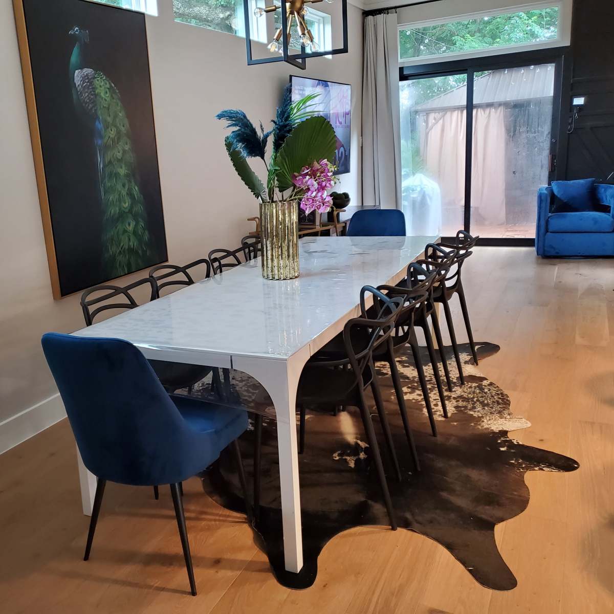 A modern dining room features a long white marble table surrounded by black chairs and a decorative vase, with a peacock painting on the wall and a blue sofa in the background.