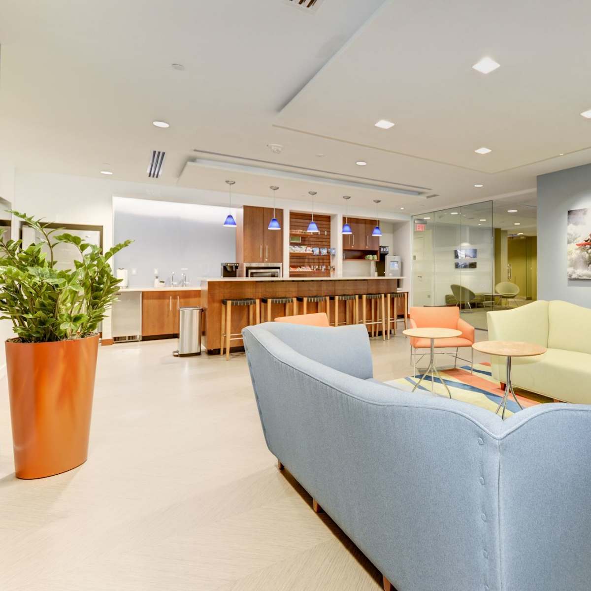 A modern lounge area featuring a blue couch, two small tables, and vibrant decor, alongside a kitchenette and a glass-walled meeting room.