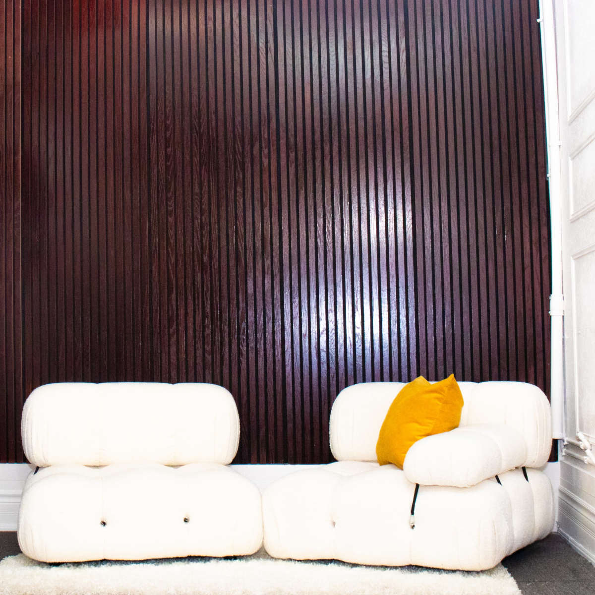 A modern, white, tufted sofa with a yellow cushion sits against a dark wooden slatted wall in a well-lit room.