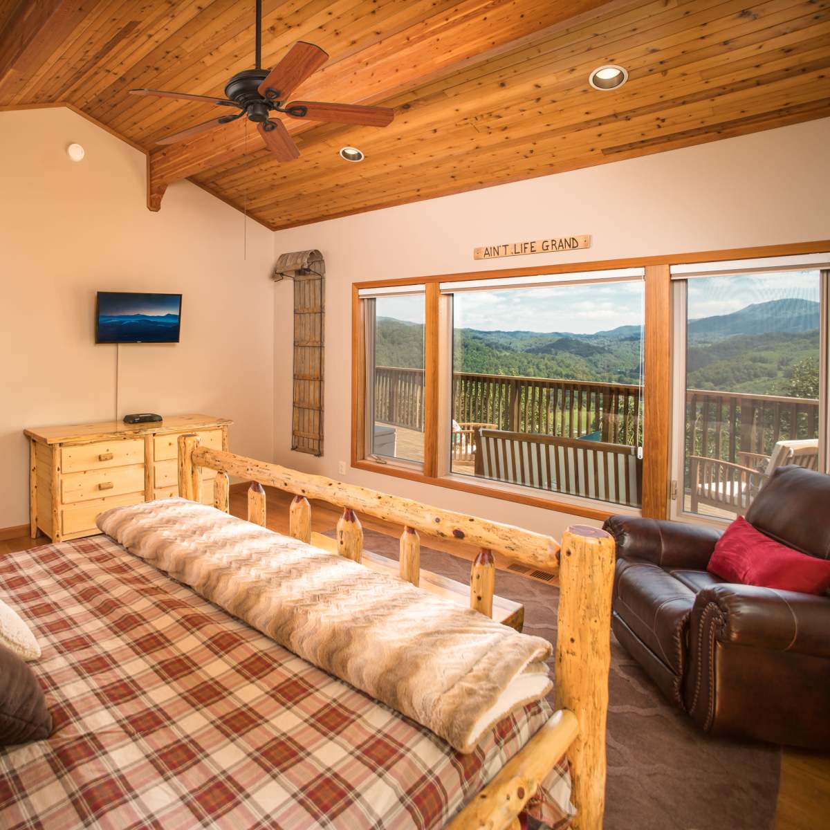 A cozy bedroom with a wooden ceiling, a log bed covered with a plaid blanket, a dark leather chair, and large windows revealing a mountain view.