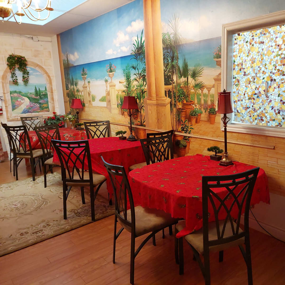 A dining area with several tables covered in red tablecloths, surrounded by decorative plants and wall murals depicting a coastal scene.