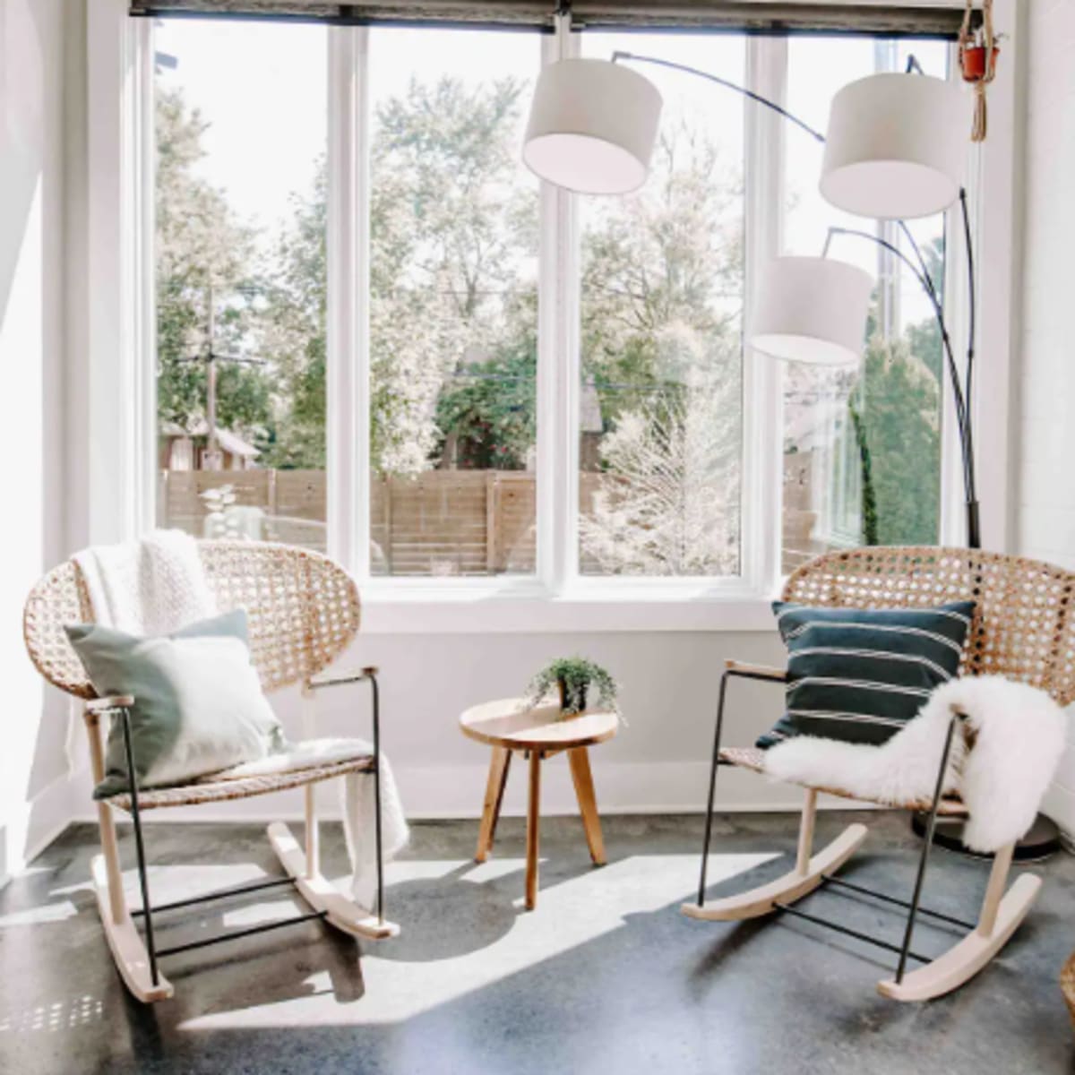 Two rocking chairs with cushions are positioned next to a small table in a sunlit corner with large windows.