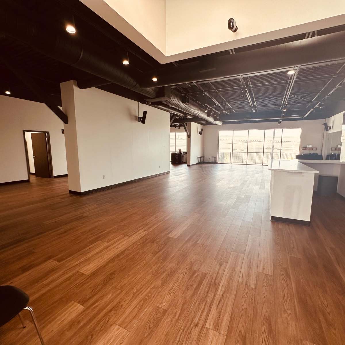 A spacious interior room with wooden floors, large windows, and a kitchenette area in the corner.