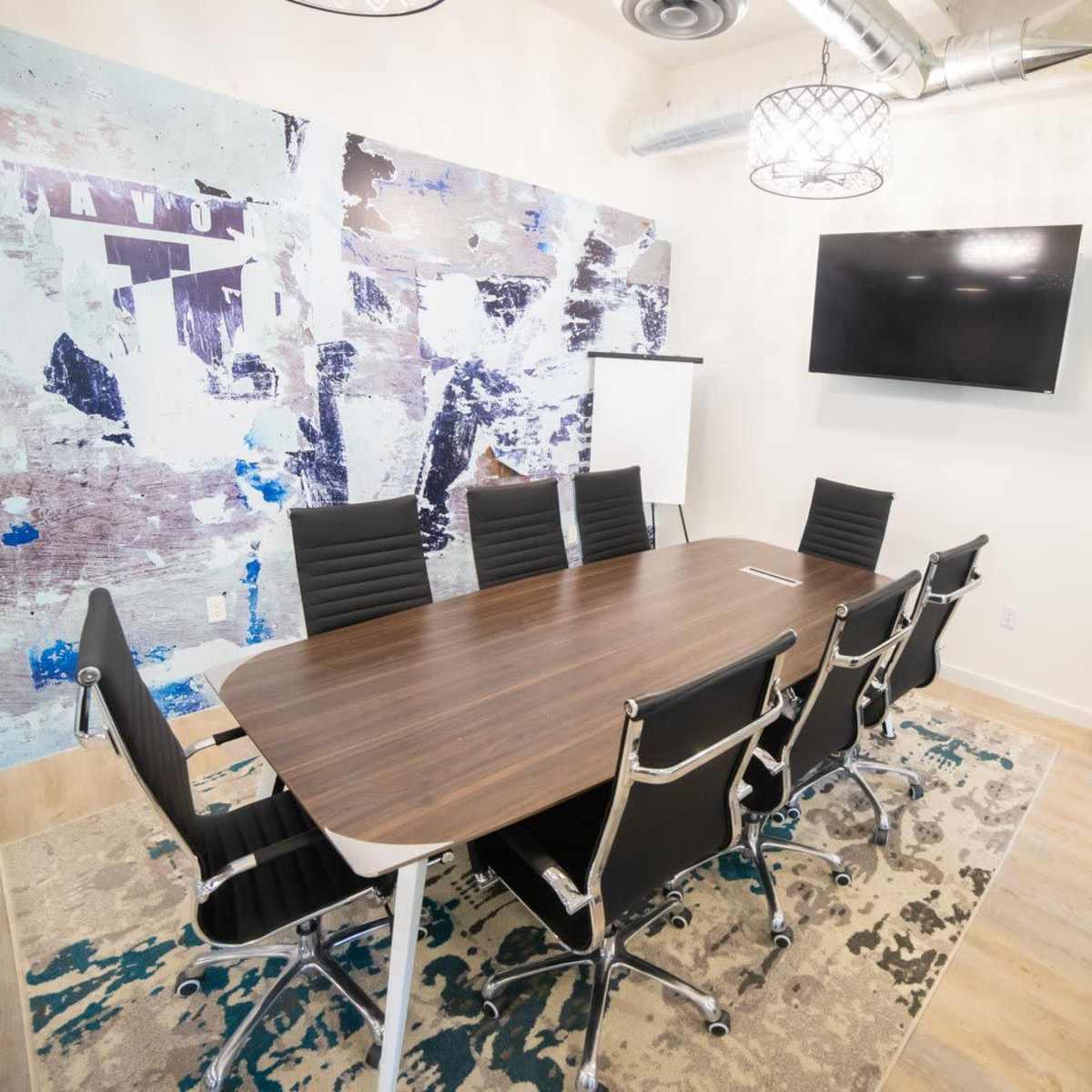 A modern conference room with a large wooden table surrounded by black chairs, a wall featuring abstract artwork, and a television mounted on the wall.
