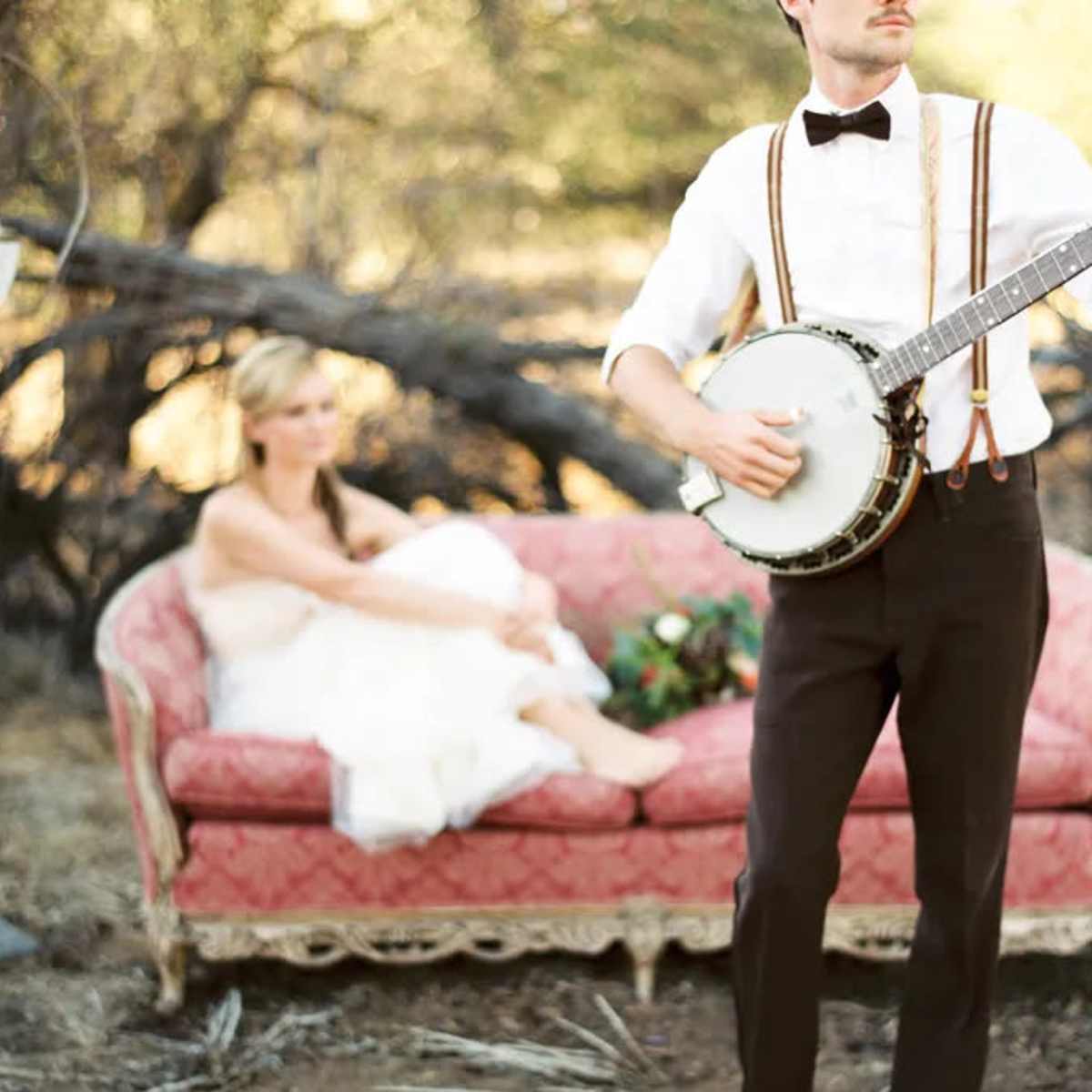 A man in formal attire plays a banjo while a woman in a white dress sits on a vintage sofa in a rural setting.