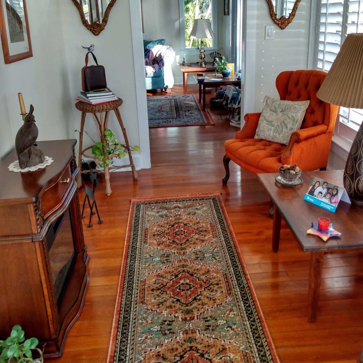 A welcoming hallway showcases a vintage rug, an orange armchair, and mirrors, leading to a bright living area with green plants and wooden furniture.